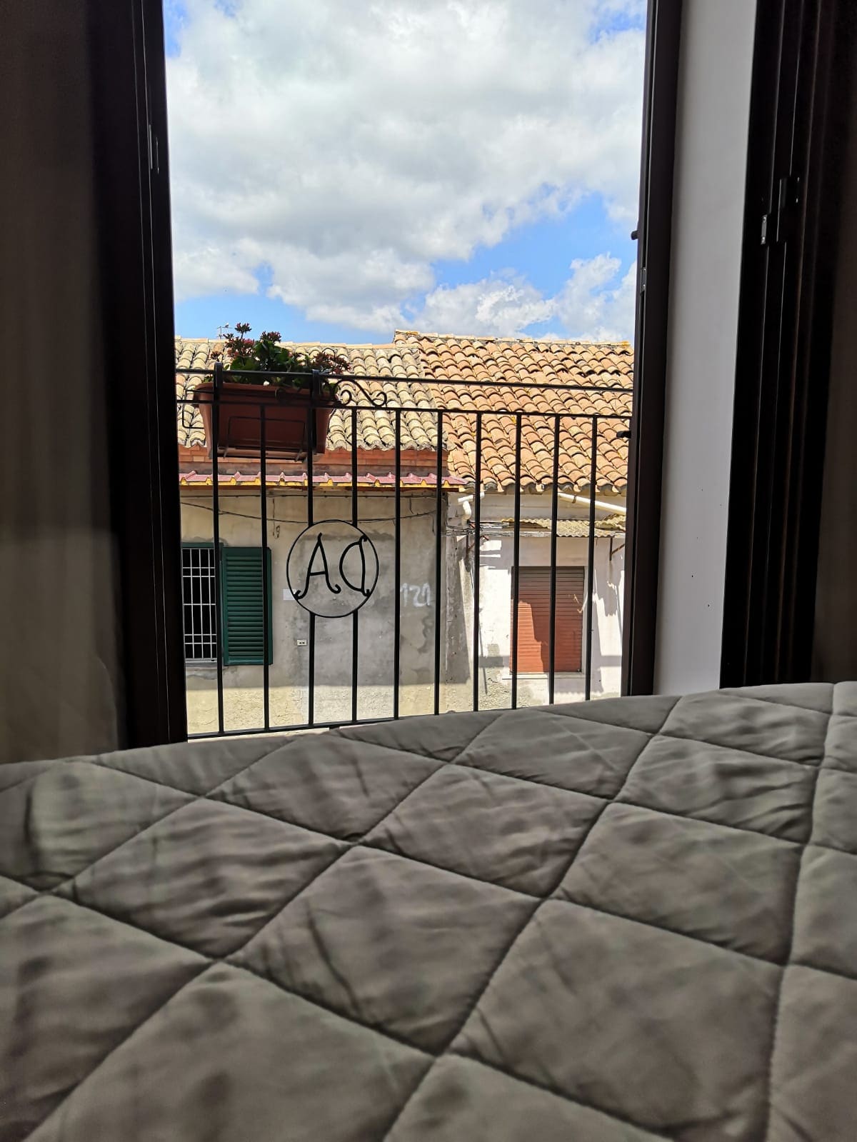 Natural light fills the room through the open window, revealing a view of neighboring buildings with tiled roofs. A decorative flower pot hangs from the wrought iron balcony, and the sky is dotted with fluffy clouds.