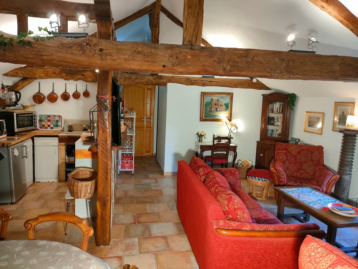 A cozy living space is visible, featuring red sofas arranged around a wooden coffee table. A dining area is seen to the left with a round table. The kitchen is equipped with modern appliances and rustic cookware displayed on the wall. Exposed wooden beams add character to the room.