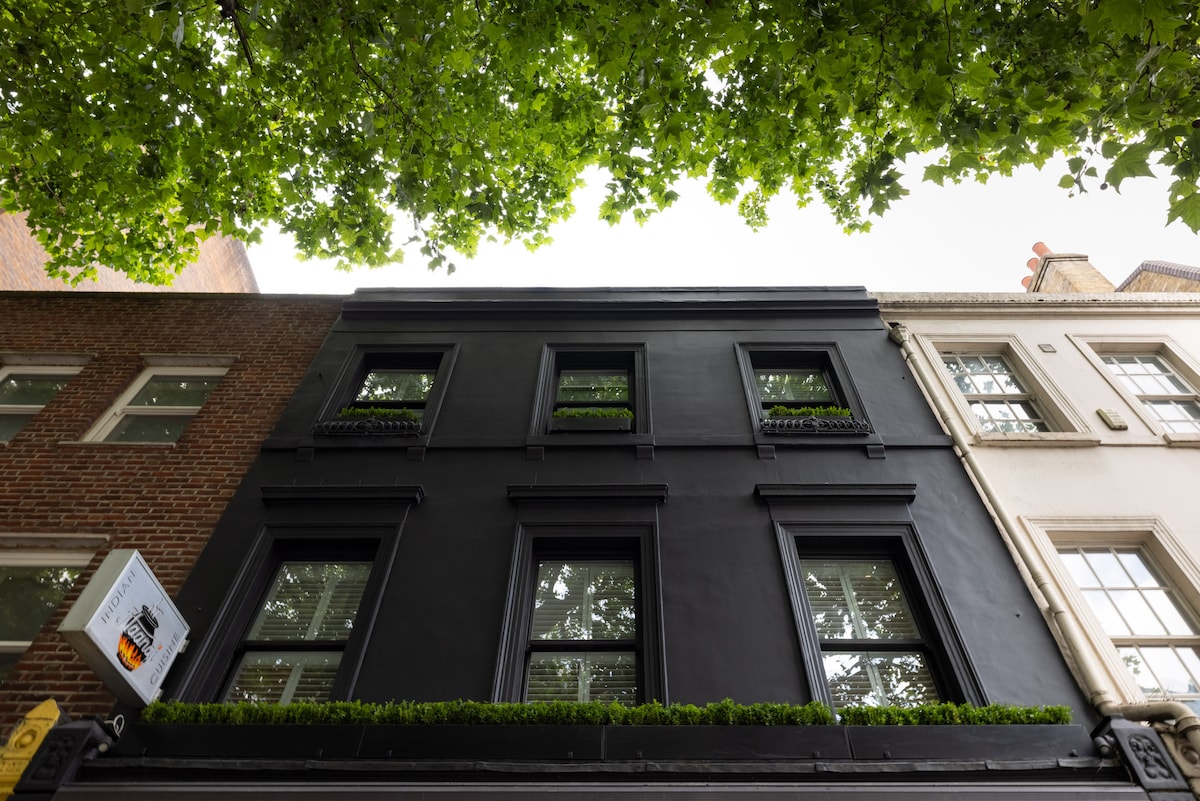 The exterior of the apartment building is viewed from below, featuring a sleek black facade and four windows adorned with small planters. Lush green leaves from a nearby tree frame the top of the image, creating a contrast against the structure.