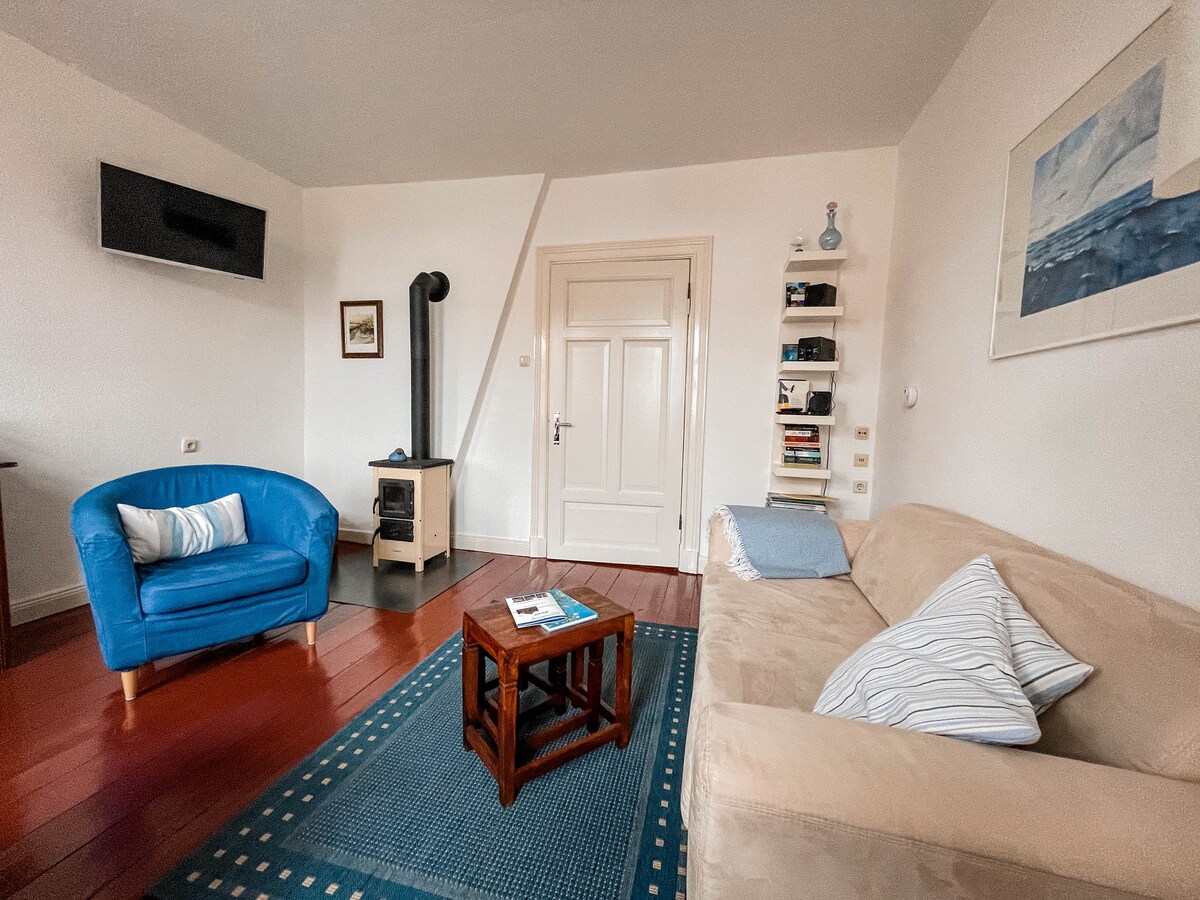 A living area is presented with a light-colored sofa and a blue armchair. A small wooden coffee table is centered on a textured area rug. A television is mounted on the wall, and a decorative shelf adds interest next to the door, leading to other areas.
