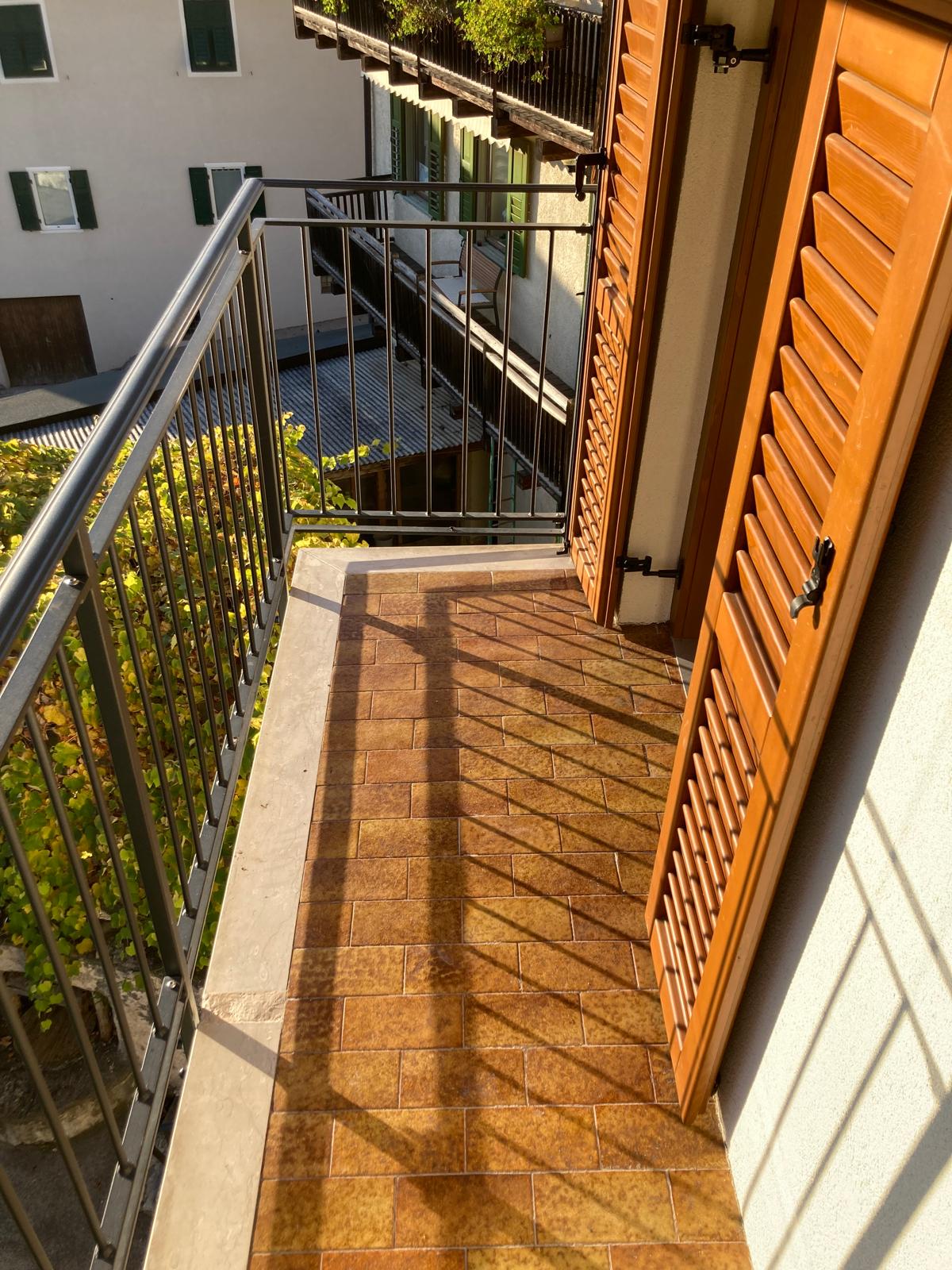 A private balcony area is depicted, featuring a textured tiled floor. Wooden shutters are visible, partially opened, allowing natural light to create patterns on the surface. The railing offers a view of neighboring buildings and greenery, enhancing the sense of outdoor space.