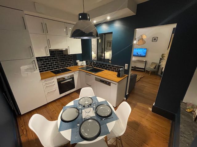 A modern kitchen area is highlighted by sleek white cabinetry and dark tile accents. A wooden countertop contrasts with the cabinetry. A round dining table is set for four with blue and white plates, surrounded by four contemporary white chairs.