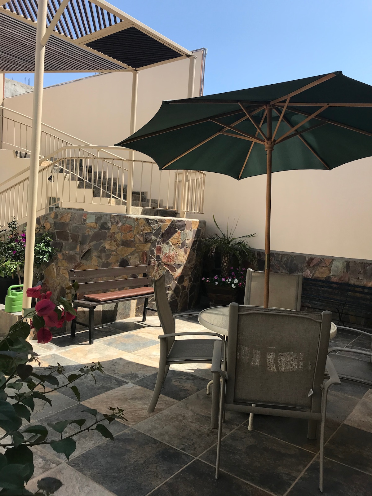A private outdoor patio is furnished with a round table and six chairs beneath a green umbrella. Stone walls adorned with potted plants surround the area, creating a serene space. A staircase leads to the upper level, and sunlight brightens the entire setting.