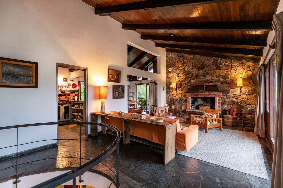 A warm living area features a stone fireplace as the focal point, framed by large windows that let in natural light. Wooden beams grace the ceiling, while a dining table and comfortable seating create an inviting space for relaxation and gatherings.