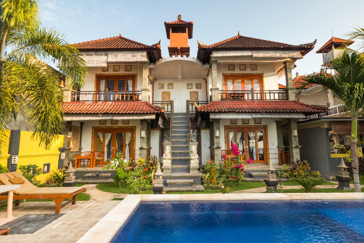 A stunning resort exterior features two-story traditional Balinese architecture with intricate carvings. Lush greenery surrounds the property, enhancing the tranquil environment. A large swimming pool reflects the clear blue sky, while spacious balconies provide views of the garden and pool area.