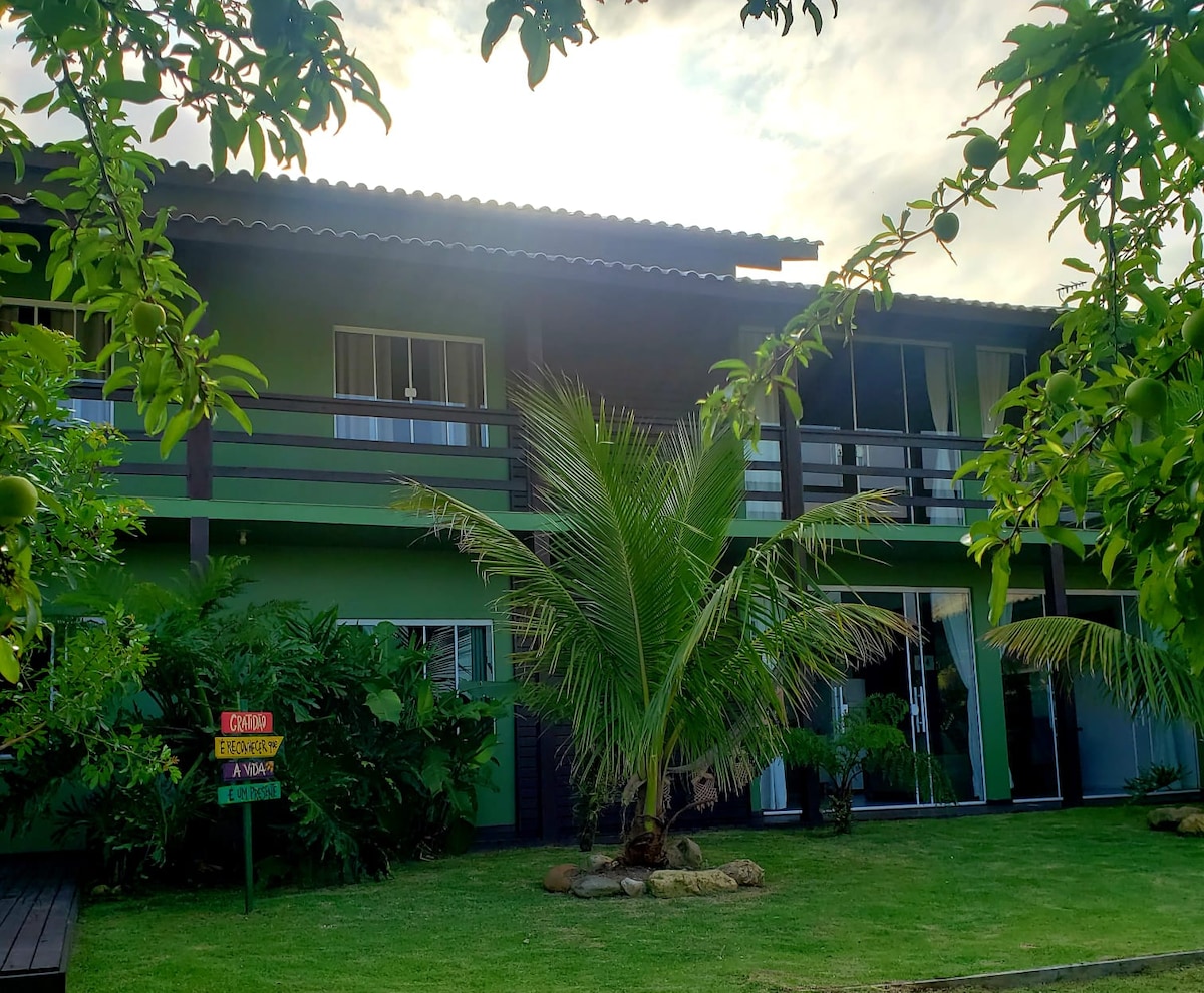 The exterior of the residence features a two-story building framed by lush greenery. A palm tree stands prominently in the foreground, with large windows allowing natural light to fill the interior spaces. The inviting balcony adds a distinctive touch to the overall design.