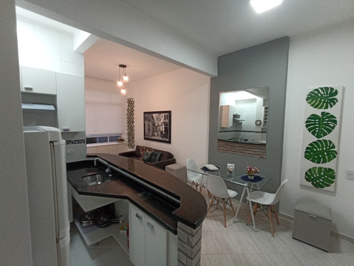 The image captures an open-plan kitchen and living area. Light-colored cabinets and a black countertop define the kitchen space, while a cozy seating area features a dark couch. A dining table with white chairs is positioned near a large mirror and wall decor with green leaf motifs.