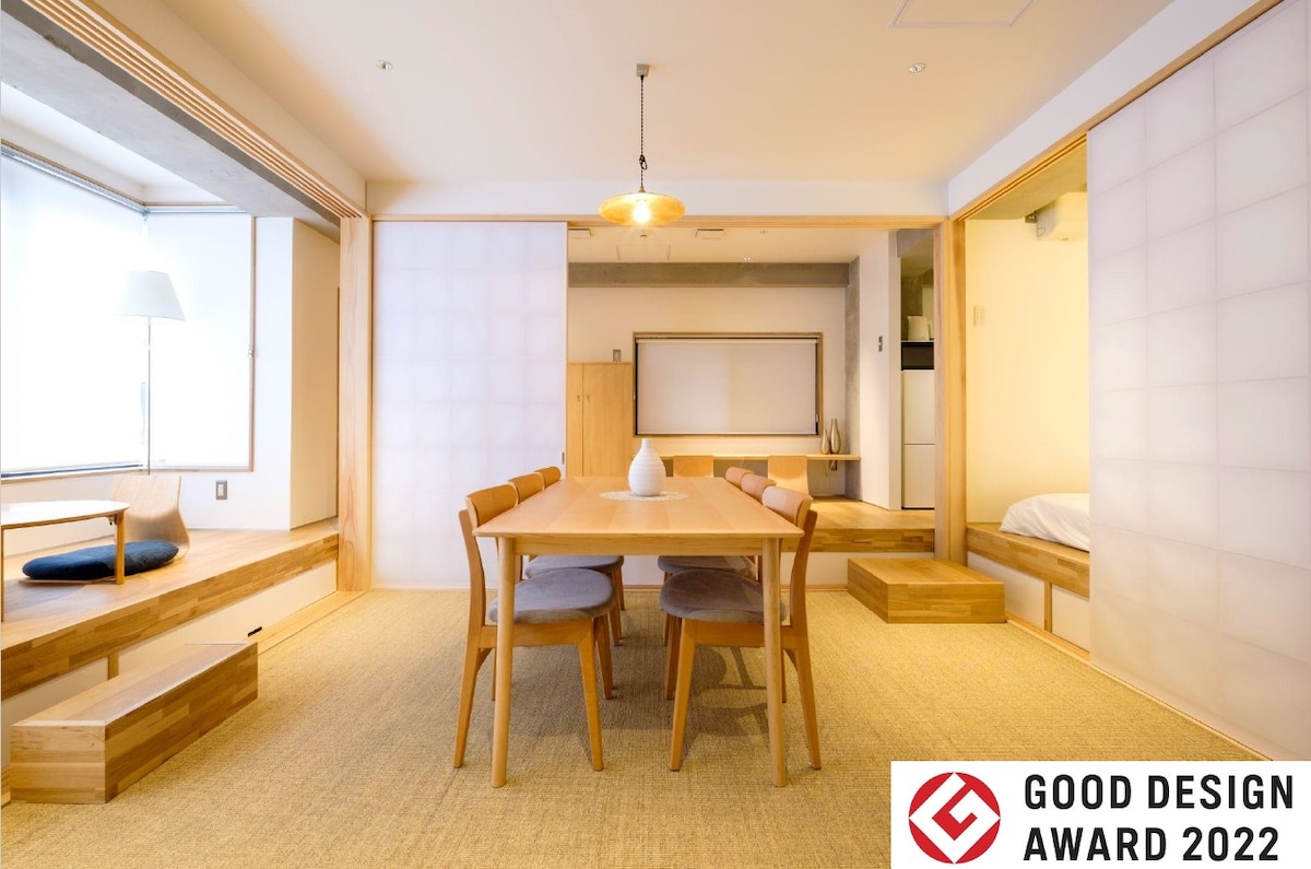 A spacious dining area features a wooden table surrounded by six chairs. Shoji doors separate the room, with seating areas visible near large windows. A neutral color palette and light wood elements create a calm environment, complemented by a soft texture from the floor.