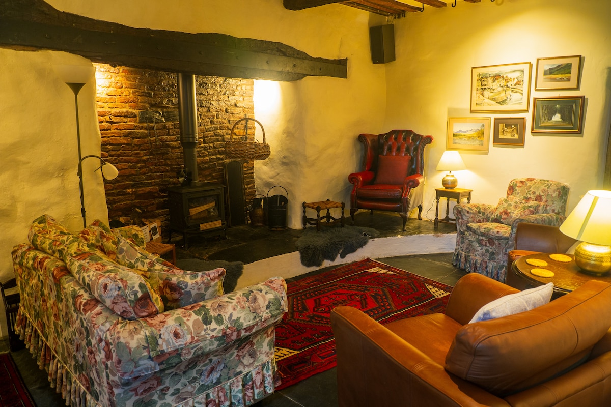 A cozy living area is displayed with floral-patterned sofas and a brown leather armchair. A well-lit fireplace made of exposed brick adds warmth, while artworks hang on the walls. A subtle area rug complements the rustic ambiance, inviting relaxation and conversation.