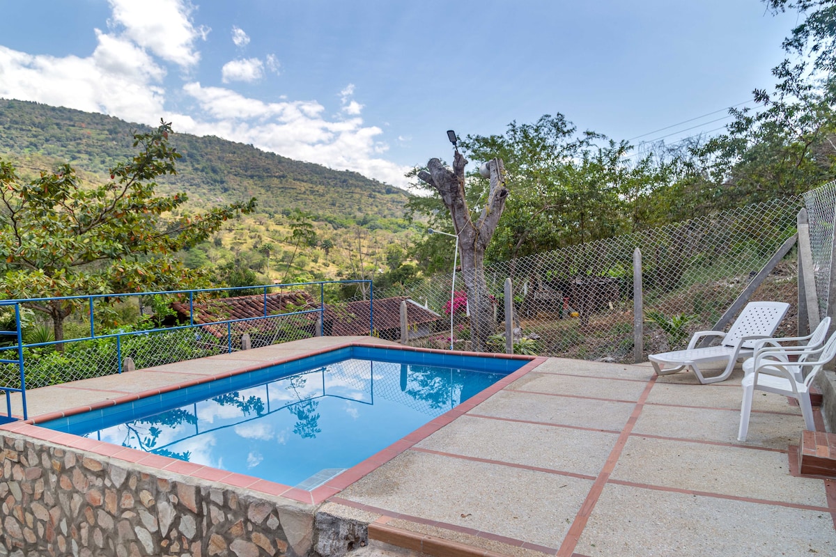 A refreshing outdoor pool is framed by a stone patio, with lounge chairs positioned nearby. Lush greenery surrounds the area, offering a serene view of the hillside, while the clear blue sky and clouds create a calm atmosphere.