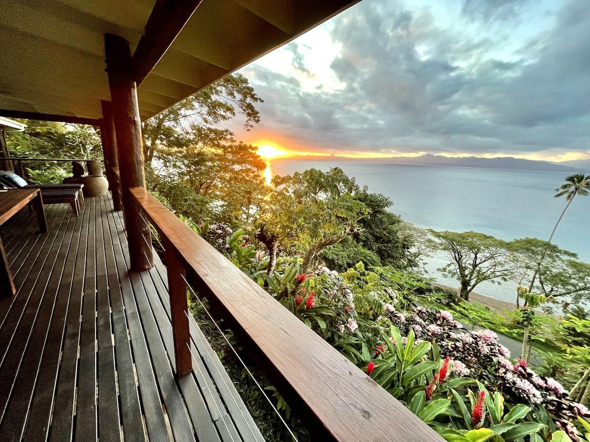 A spacious deck provides an expansive view of the ocean and sunset, framed by lush greenery and colorful flowers. The wooden railing enhances the natural aesthetics, creating a seamless connection with the surrounding landscape.