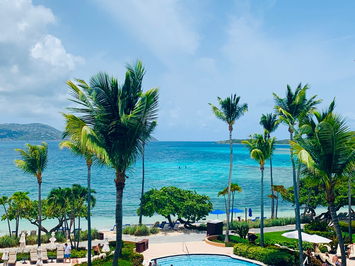 A serene beachfront view captures the turquoise ocean and gentle waves lapping at the shore. Lush palm trees frame the scene, while a sparkling pool is visible in the foreground. The horizon reveals distant islands beneath a clear blue sky.