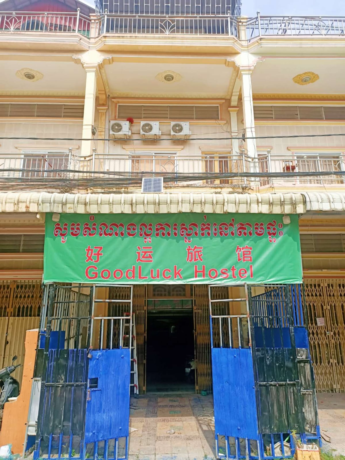 The exterior of the hostel is characterized by a large, vivid green sign that displays the name 'Good Luck Hostel' in both English and Chinese. The building features multiple floors with balconies, and large air conditioning units are mounted on the wall.