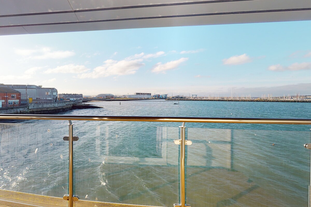 A private balcony is viewed from inside, featuring a glass railing that allows for unobstructed views of Portland Harbour. Calm waters reflect the sky, with the shoreline and infrastructure visible in the distance, showcasing the proximity to coastal landscapes.