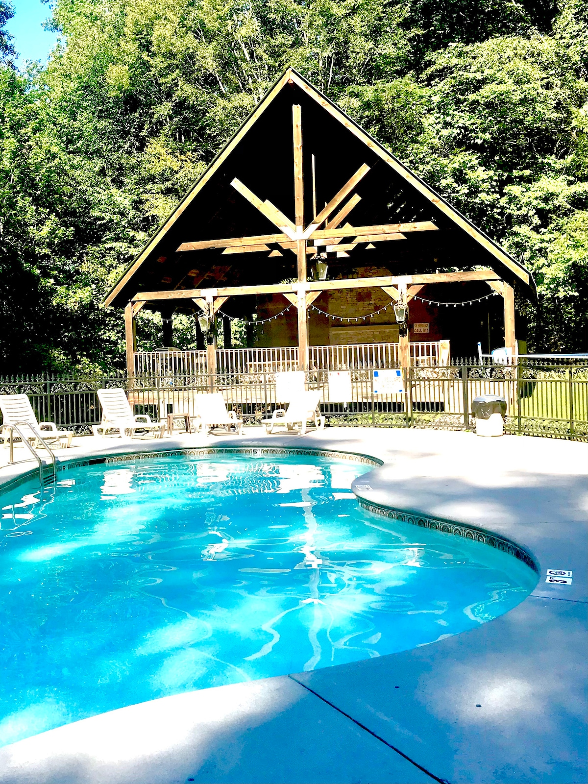 A swimming pool is surrounded by a concrete deck with several lounge chairs. A covered pavilion is visible in the background, characterized by a peaked roof and wooden beams, providing shaded seating for guests near the pool area.
