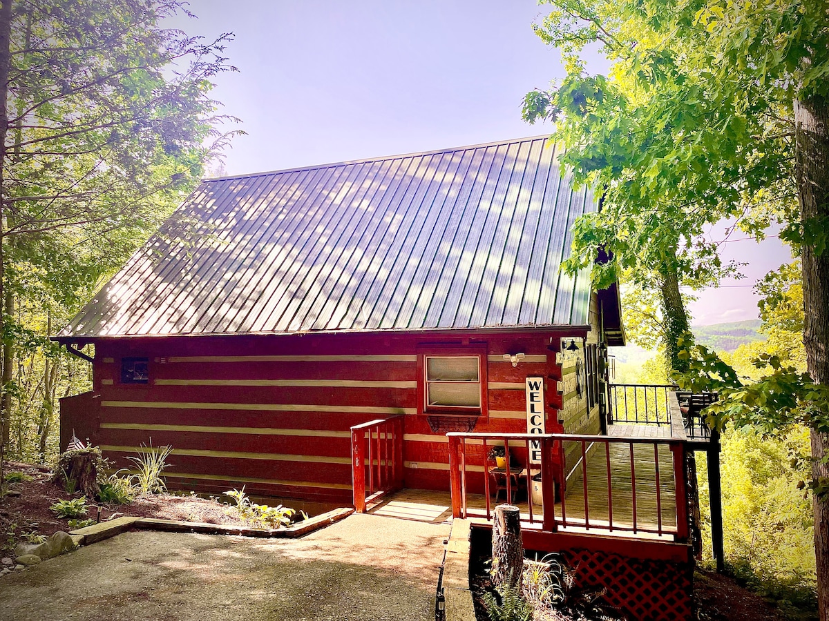 The cabin features a rustic exterior with wooden logs and a metal roof, surrounded by lush greenery. A welcoming sign is visible by the entrance, which leads to a spacious deck. Natural light highlights the inviting atmosphere of this secluded retreat.