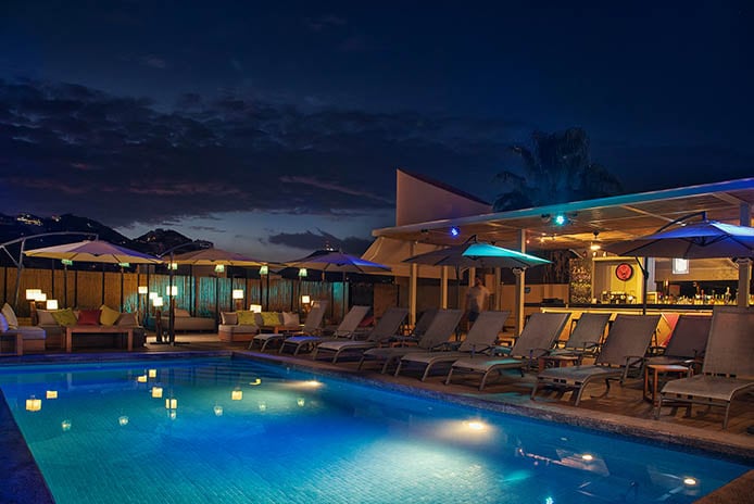 A tranquil pool area is illuminated by soft lights under a night sky. Sun loungers with umbrellas are arranged around the pool, creating inviting spots for relaxation. A bar area is visible in the background, enhancing the communal atmosphere.