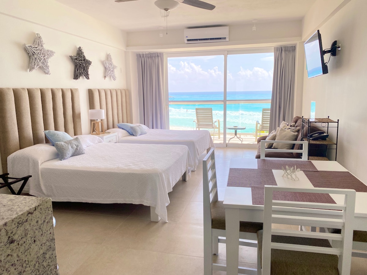 The studio features two spacious beds with neutral bedding, alongside a dining area with a table and chairs. Large glass doors provide a view of the beach and ocean, while a small seating area is present on the balcony, enhancing the connection to the outdoor scenery.