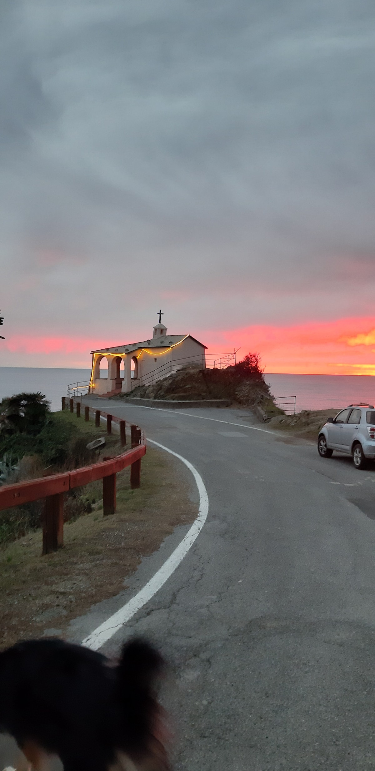 A narrow road leads to a small chapel, illuminated against a vibrant sunset sky. The scene is framed by a wooden railing on one side and a parked car nearby. The surrounding landscape features gentle slopes and greenery.