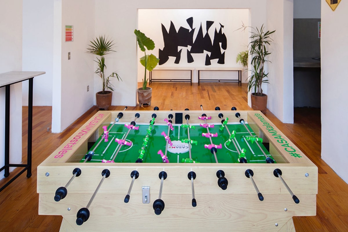 A foosball table with colorful players is prominently displayed in a spacious common area. Bright greenery from potted plants complements the artistic black-and-white wall piece visible in the background. The warm wooden floor enhances the inviting atmosphere of the space.