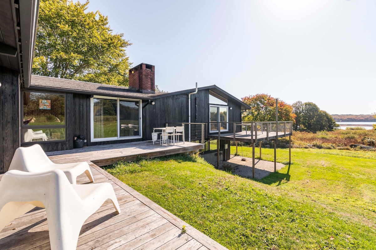An outdoor view of a midcentury house featuring large windows and a spacious deck. A dining table with chairs is positioned on the deck, overlooking a lush green lawn and scenic landscape. The setting is surrounded by trees displaying autumn colors.
