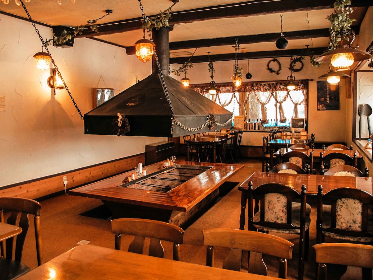 A spacious dining area is presented with wooden tables and chairs arranged around a centrally located grill under a hood. Soft lighting emanates from hanging lamps, enhancing a warm atmosphere. Large windows are adorned with curtains, allowing natural light to filter in.