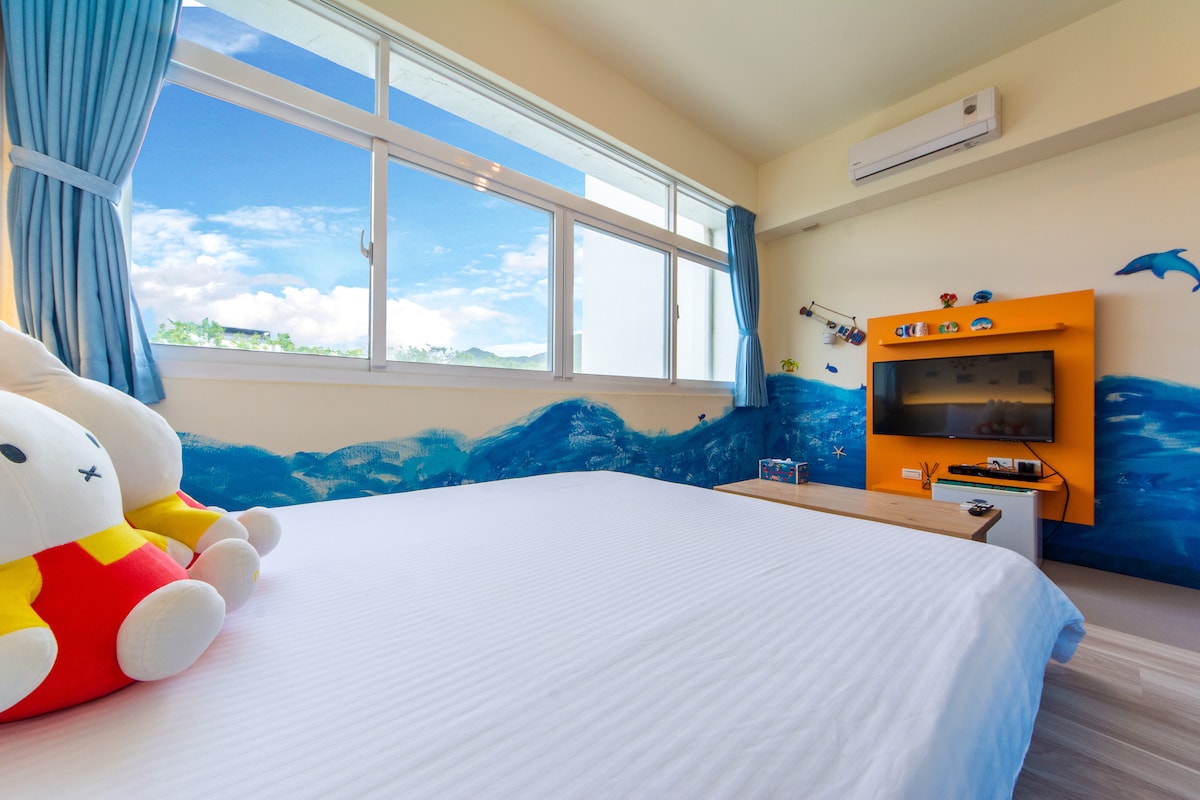 A bright bedroom features a spacious bed with a neatly arranged white comforter. A large window provides a view of the blue sky, while cheerful wall decor of waves and dolphins enhances the playful atmosphere. A TV is available on an orange cabinet.