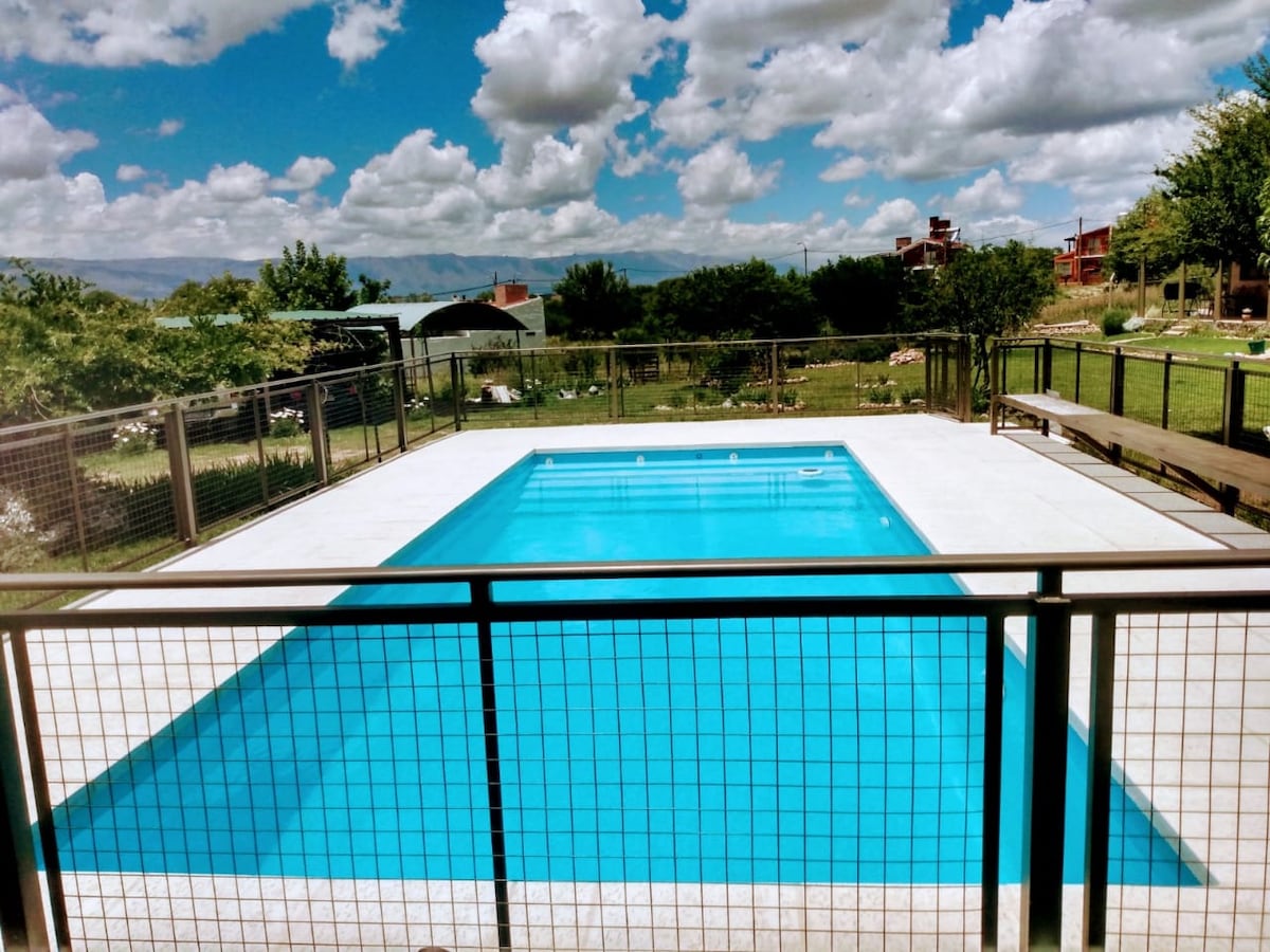A clear swimming pool is surrounded by a fenced area, featuring a spacious deck. Green grass and trees create a natural backdrop, with fluffy clouds scattered in the blue sky above.