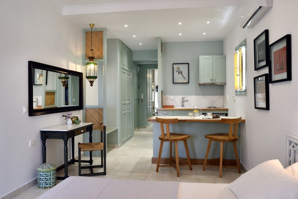 An open-plan studio apartment features two single beds and a small dining area, flanked by a kitchenette. A mirror and lantern provide soft lighting, while natural light enters through the windows. Light-colored walls complement the wooden furniture, creating a calm and welcoming environment.