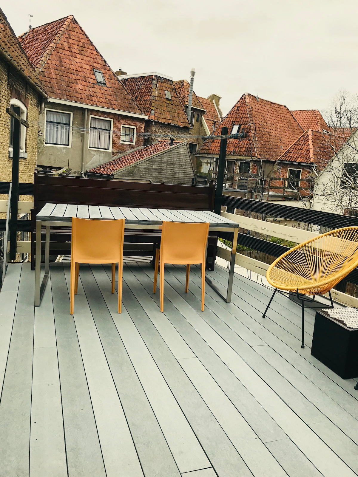 An outdoor terrace is shown with a wooden deck and a modern grey table accompanied by two yellow chairs. The backdrop features sloped roofs of neighboring buildings, creating a quaint urban environment. A cozy chair is positioned nearby, enhancing the seating arrangement.