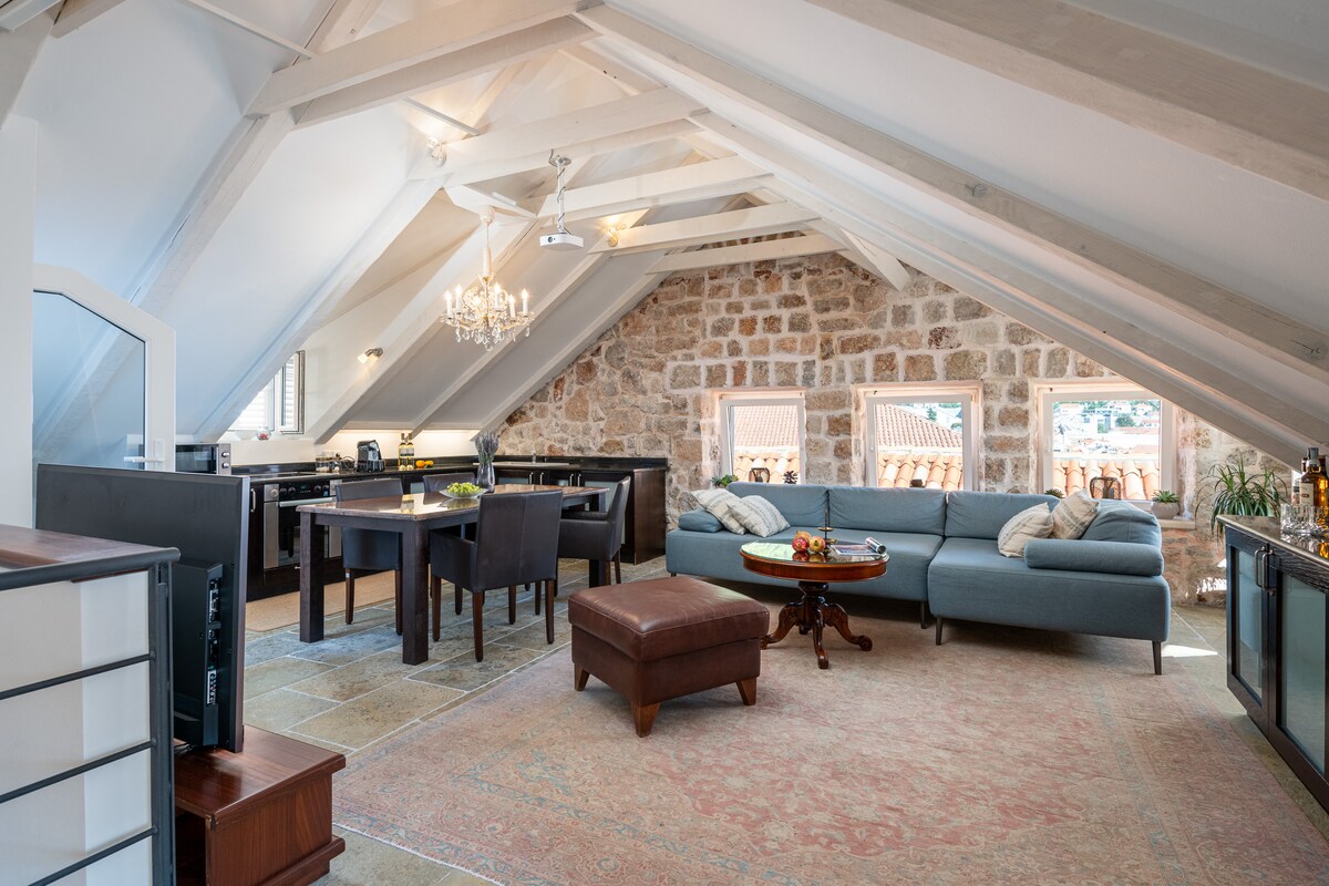 The spacious living area features a high-pitched ceiling with exposed wooden beams. A comfortable blue sofa and a wooden dining table occupy the center, while large windows offer views of the surroundings. A decorative chandelier adds a touch of elegance to the space.