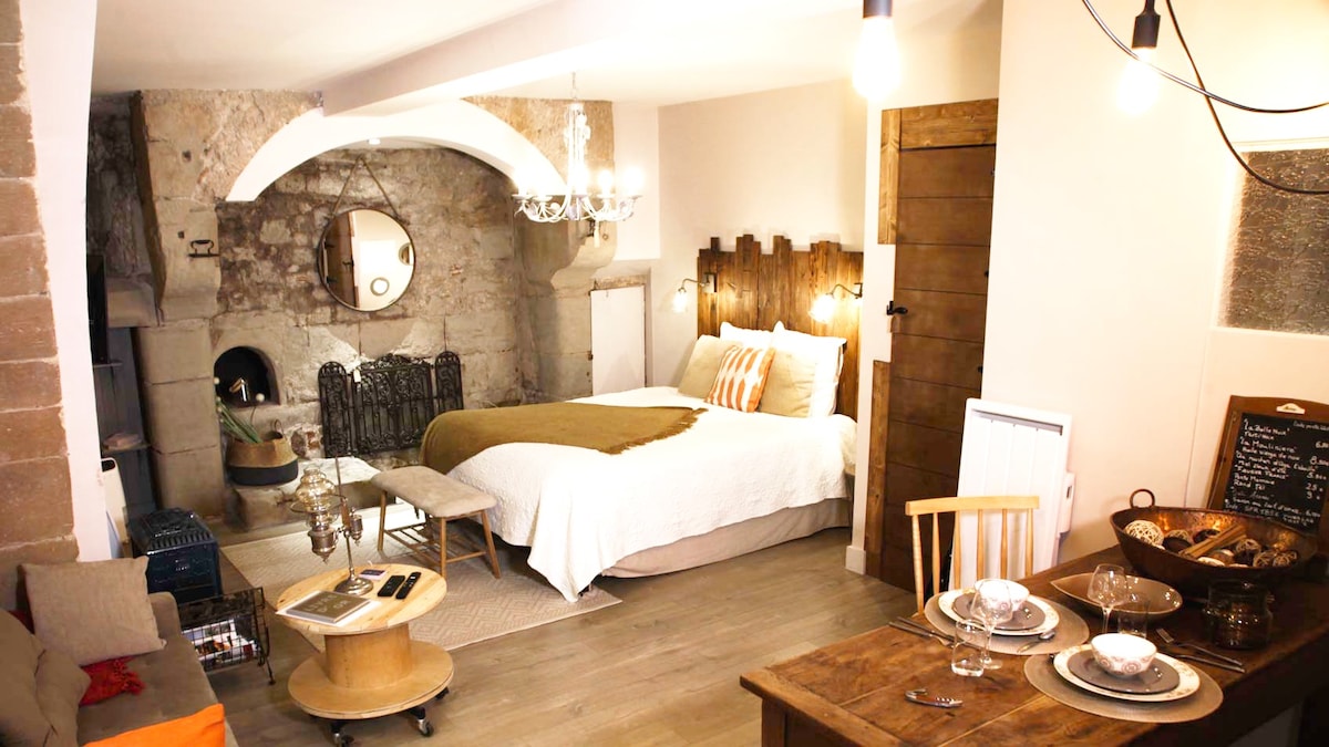 A studio space features a spacious bed with a neutral-colored blanket, complemented by warm light fixtures. A vintage stone wall serves as a backdrop, next to an elegant chandelier. A wooden dining table is set for two, inviting relaxation and comfort.