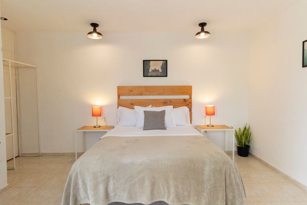 A spacious room is shown with a large bed featuring a wooden headboard. Soft lighting is provided by two bedside lamps. A small plant is positioned near the floor, and a closet is visible in the background. Neutral tones create a calming atmosphere.