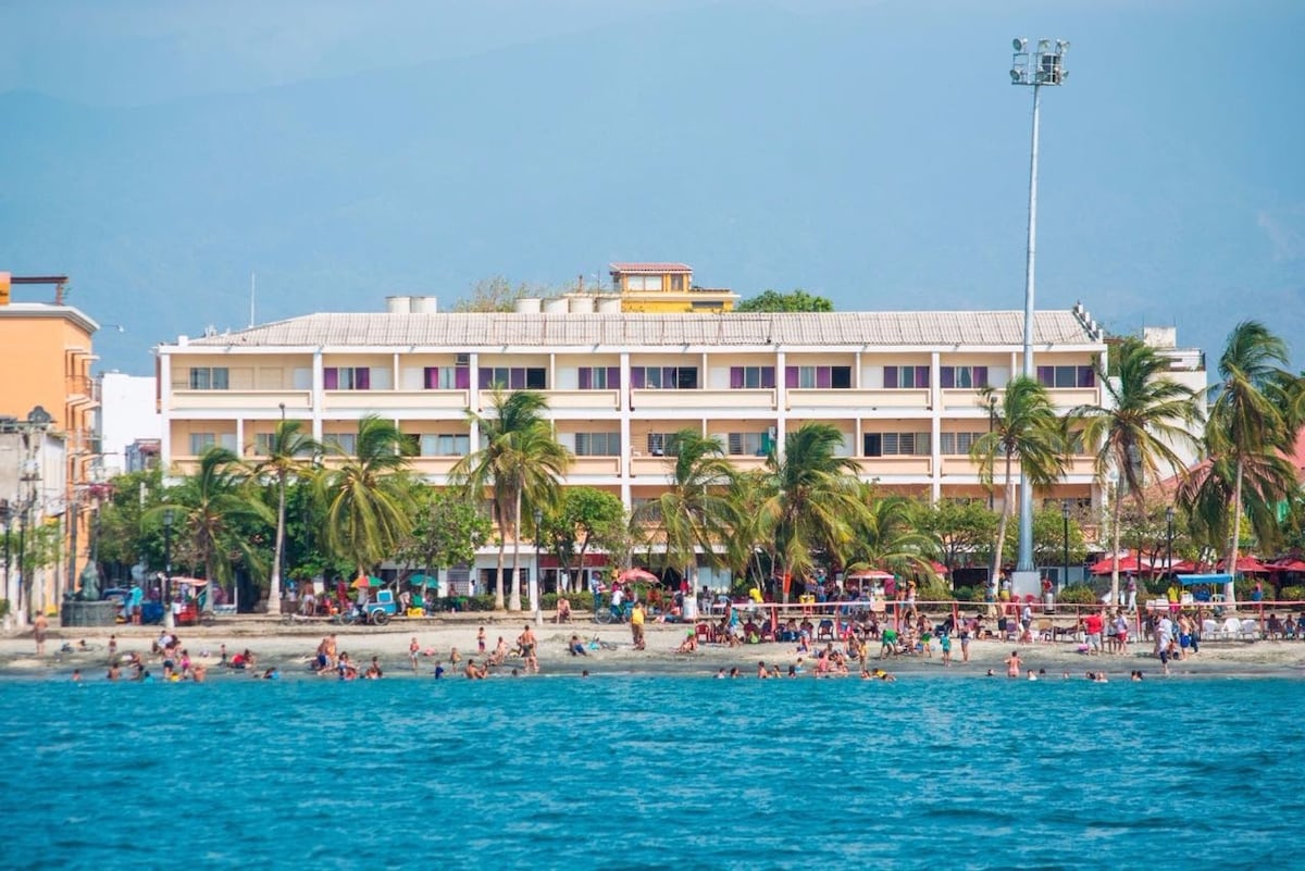 A beachfront view is presented, highlighting a three-story building surrounded by palm trees. The vibrant beach is populated with visitors enjoying the water, while colorful umbrellas and loungers are positioned along the shore. Mountains loom in the background under a clear sky.