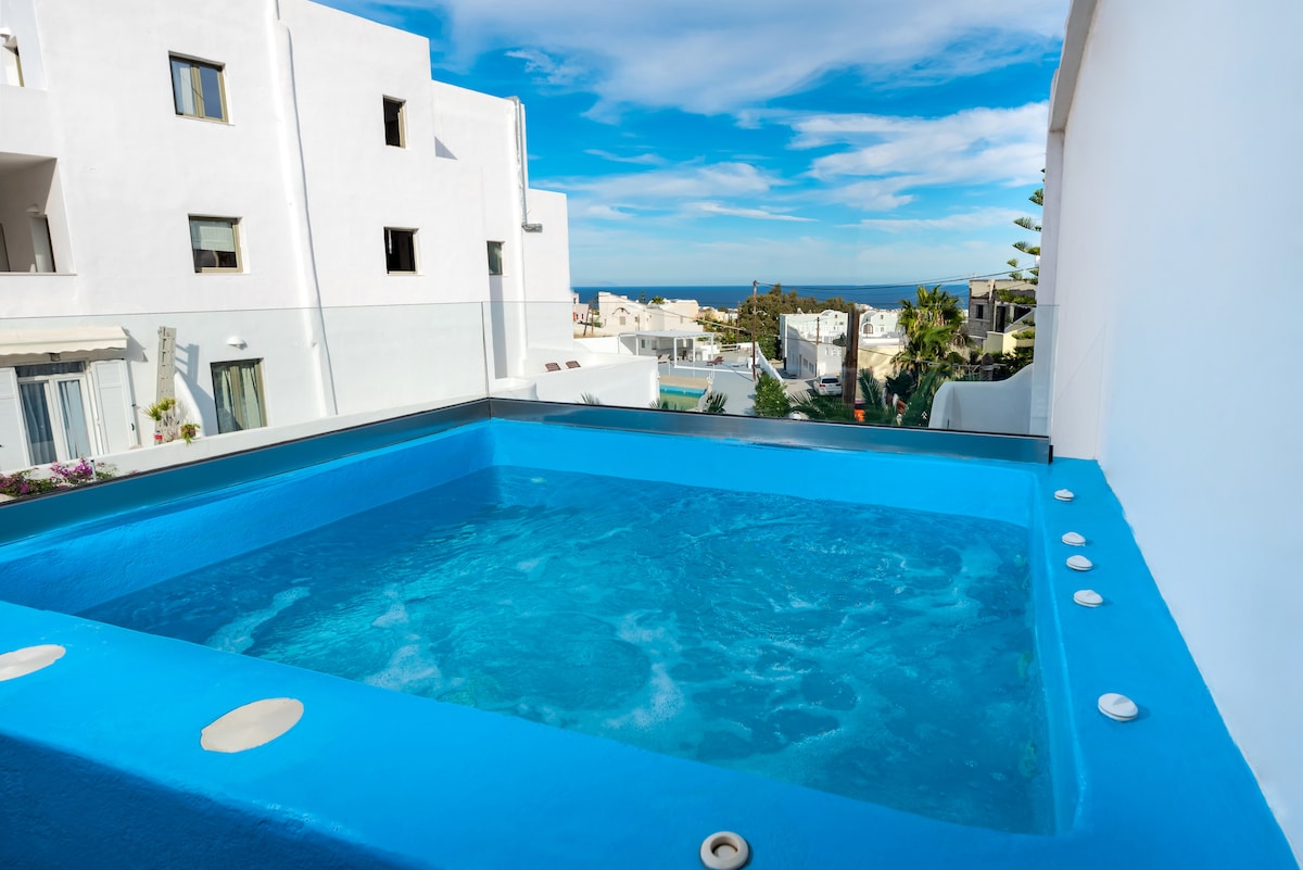 A heated outdoor jacuzzi is situated on a private balcony, surrounded by colorful landscapes and offering a clear view of the sea in the distance. The bright blue water invites relaxation and comfort, framed by modern white buildings.