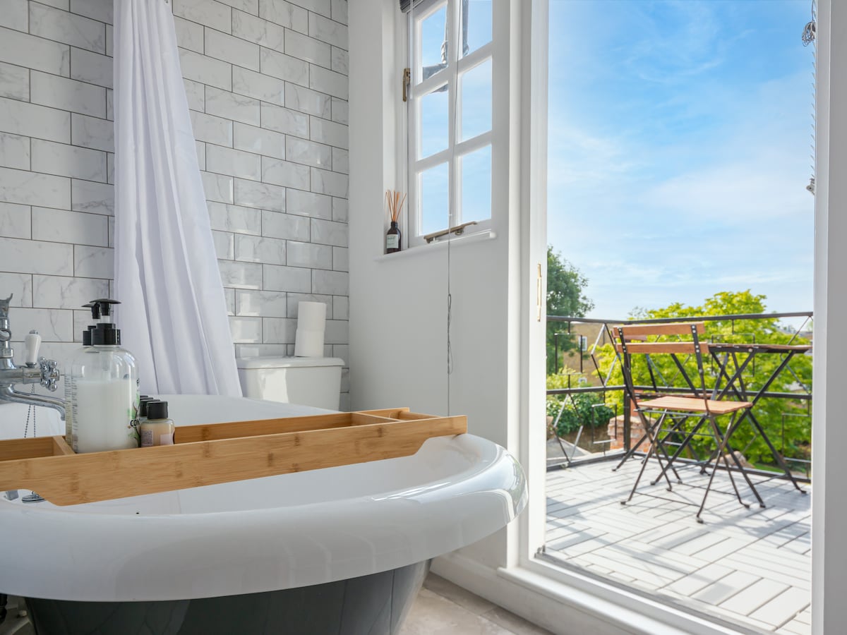 A bright bathroom features a luxurious roll-top bath with a wooden tray displaying toiletries. Large windows provide a view of a private terrace with a small dining set for two, overlooking greenery and blue skies.