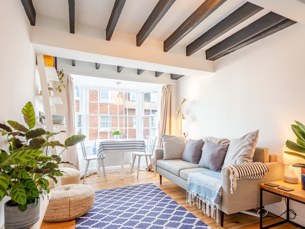 A stylish living area features a comfortable sofa adorned with cushions. A round light fixture hangs above a dining table set for two. Natural light floods through large windows, creating a bright and airy environment. Indoor greenery adds a touch of nature to the space.