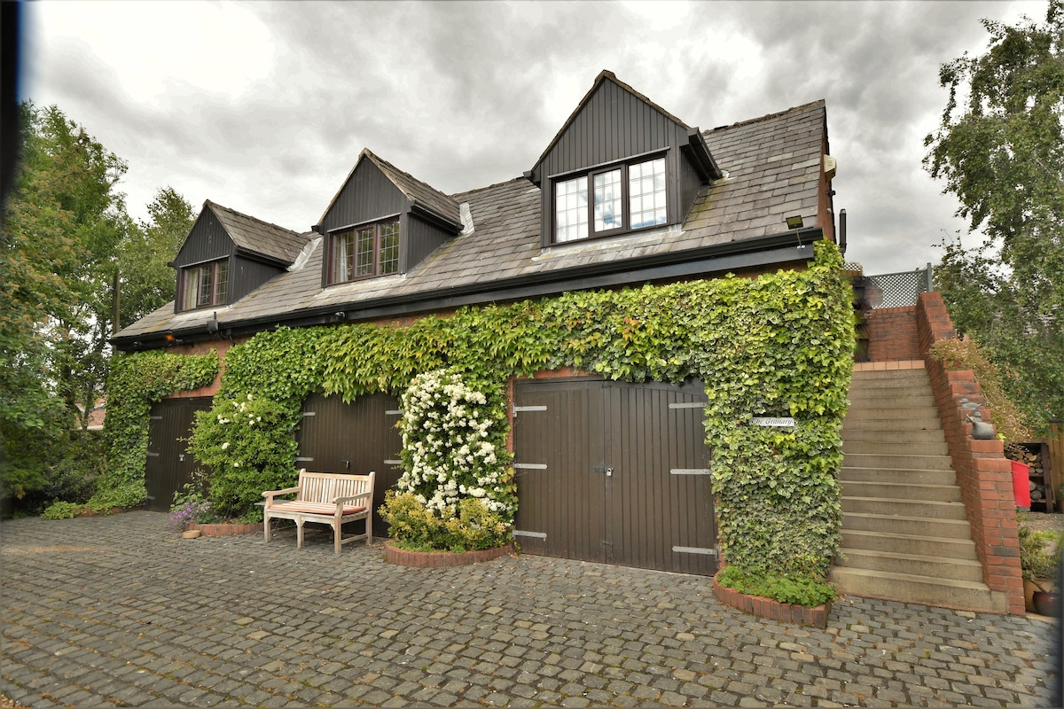 The exterior of a charming black-clad building is surrounded by lush greenery, with vines climbing the walls. The façade features multiple windows and double doors, while a cobblestone driveway provides access. A wooden bench sits along the flower-adorned walkway leading to the entrance.