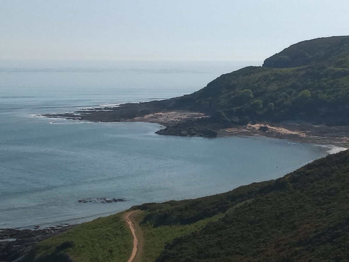 A serene coastal view captures gently sloping hills rising from a tranquil sea. The clear water reflects the sky, while a winding path meanders along the shore. Rocky outcrops are visible, with soft sandy stretches in the distance, framed by lush greenery on the hills.