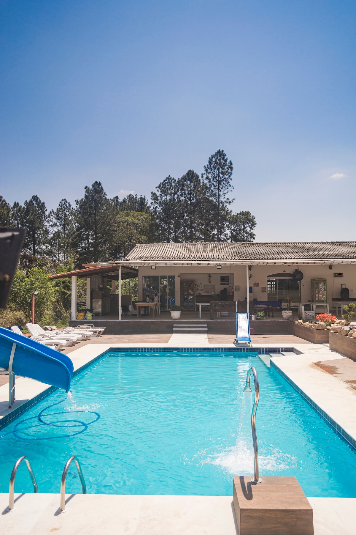 A refreshing pool area features a blue water slide, surrounded by lounge chairs under the sun. The spacious outdoor area provides access to a charming house visible in the background, with greenery framing the scene. The clear sky enhances the inviting atmosphere.