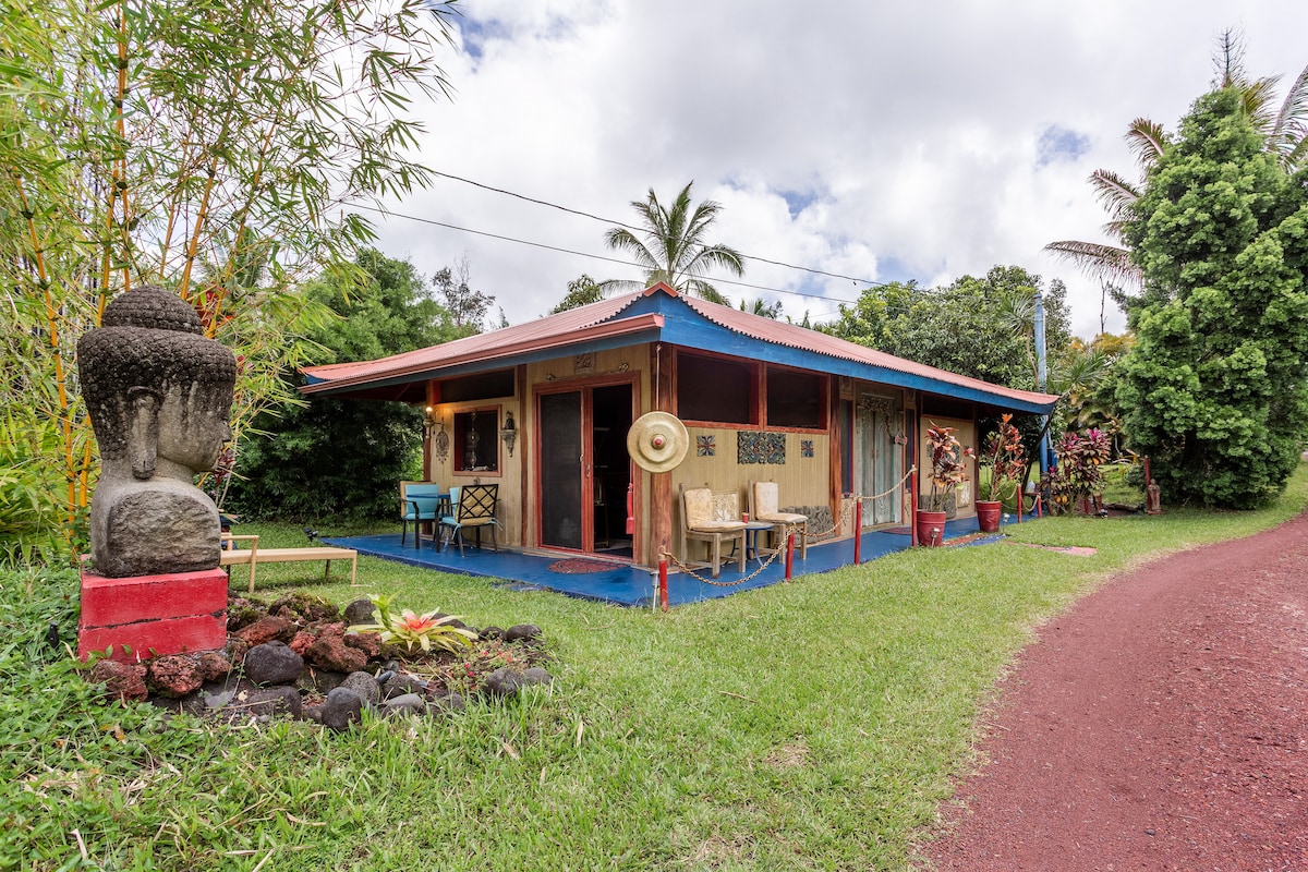 A colorful house featuring a spacious porch is surrounded by lush greenery. Statues and tropical plants enhance the inviting exterior. A red dirt path leads to the entrance, while a blue roof complements the vibrant decor, creating a harmonious blend with the natural landscape.