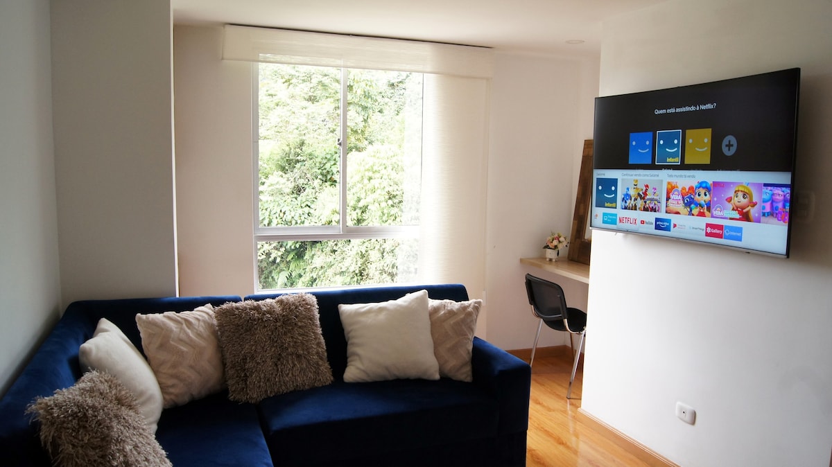 A comfortable living area features a navy blue sofa adorned with decorative cushions. A large window provides natural light and views of greenery. A wall-mounted TV displays streaming options, while a small desk and chair are positioned nearby.