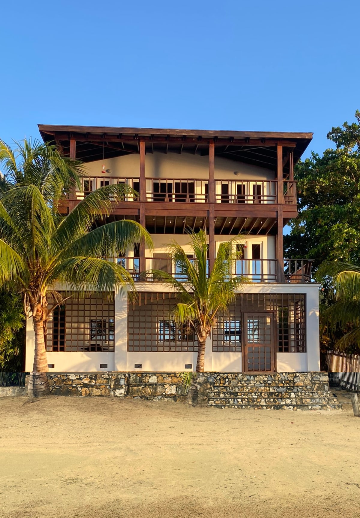 The three-story villa is framed by palm trees, showcasing a blend of wood and stucco architecture. Balconies adorn each floor, allowing access to outdoor spaces. The sandy area in front enhances the beach-like ambiance, creating a welcoming entrance to the property.