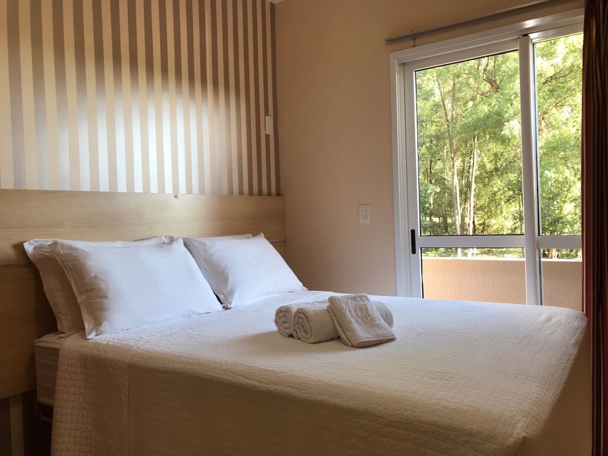 A bright bedroom features a spacious bed with neatly arranged white linens and towels. Soft light enters through a large glass door leading to a balcony, surrounded by greenery. Warm beige walls with subtle stripes provide a calming atmosphere.