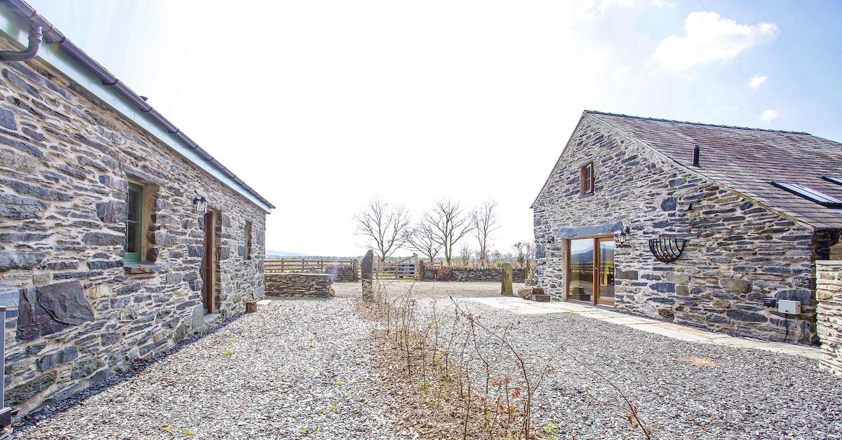 A spacious courtyard is surrounded by stone buildings, showcasing a blend of rustic charm and contemporary design. A gravel surface extends towards the entrance of the cottage, with views of the landscape and trees in the background under a bright sky.