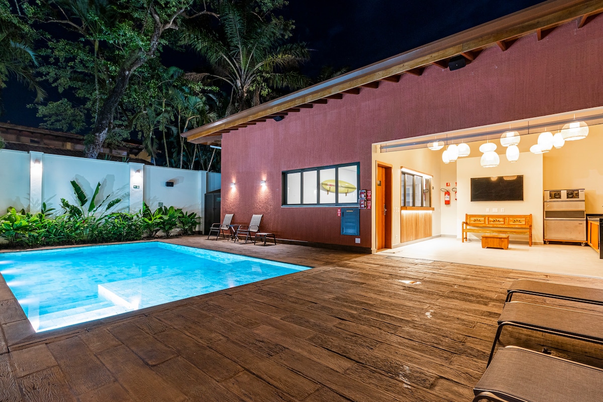 An outdoor pool area is illuminated at night, surrounded by lush greenery. Light wood decking complements the pool’s clear water, while loungers are positioned nearby. The adjoining building showcases modern lighting fixtures and large windows, offering a view of the inviting indoor space.