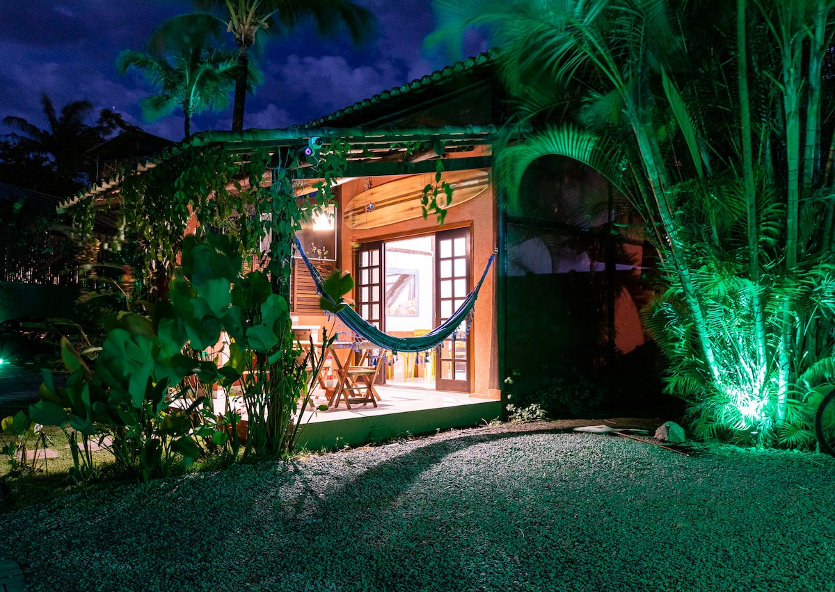 A cozy exterior view of a wooden cabin is enhanced by soft lighting from nearby palm trees. A hammock is suspended between two support beams, inviting relaxation. The entrance is framed by foliage, creating a serene atmosphere with natural elements and decorative lighting.