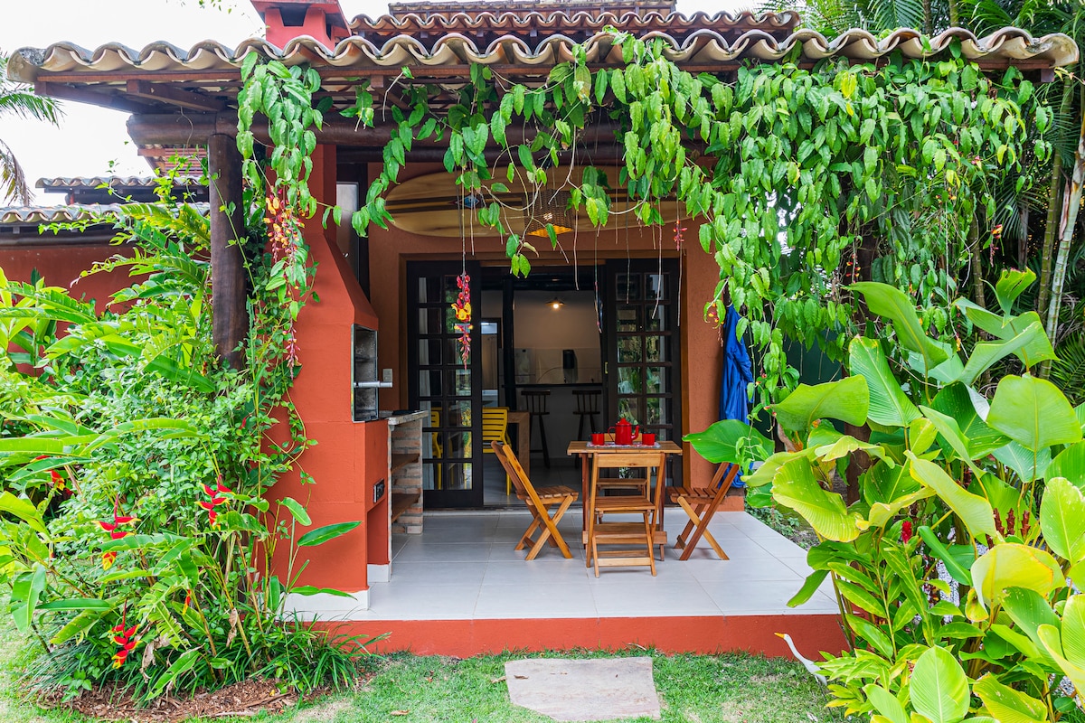 The exterior of the chalet is adorned with lush green vines and tropical plants, providing a vibrant backdrop. A small patio features a wooden dining table surrounded by four chairs, ideal for outdoor meals. A grill is located nearby, enhancing the space's functionality.