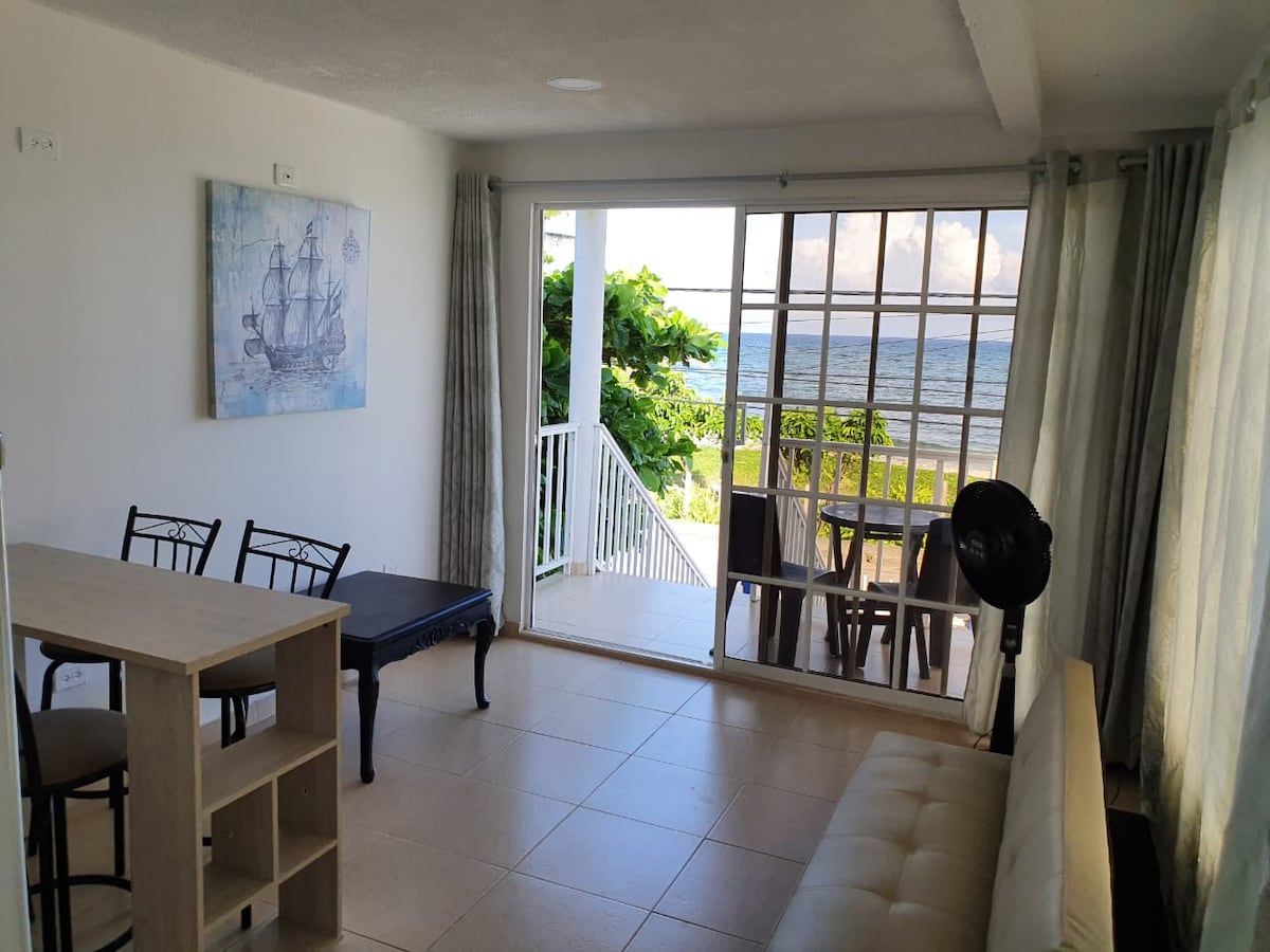 A light-filled living area features a neutral-colored sofa, a small dining table with chairs, and a wall-mounted piece of artwork depicting a ship. Large sliding glass doors open towards a scenic view of the beach and outdoor seating on a balcony.