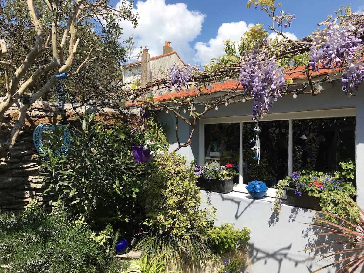 A charming outdoor patio is adorned with flowering plants and greenery. Brightly colored pots and hanging decorations enhance the natural beauty, while a clear window showcases the inviting interior. Decorative string lights are interwoven among the foliage, adding a touch of whimsy to the serene space.